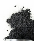 A close-up of a pile of fine black powder spilled onto a white surface, with the edge of a small container visible at the bottom of the image.
