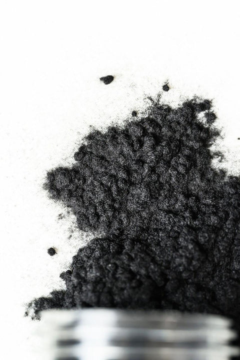 A close-up of a pile of fine black powder spilled onto a white surface, with the edge of a small container visible at the bottom of the image.