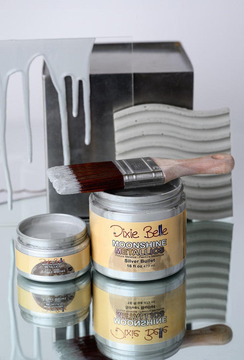 Two open jars of Dixie Belle Moonshine Metallics paint, one large and one small, with a paintbrush resting on the larger jar. Silver paint drips and painted surfaces are visible in the background.