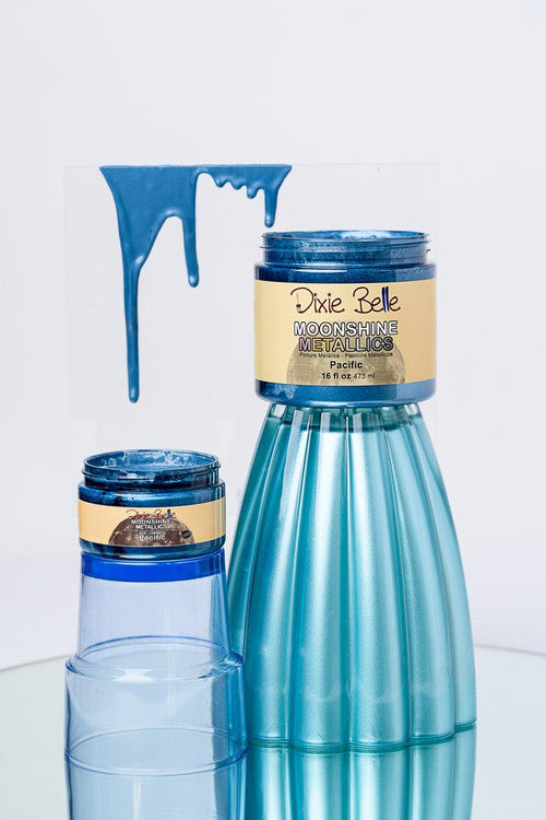 Two containers of Dixie Belle Moonshine Metallics paint in Pacific blue sit on a reflective surface. Showing paint dripping from a clear sheet above.