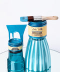 Two blue metallic paint jars, one large and one small, labeled "Dixie Belle Moonshine Metallics Caribbean," are on a mirrored surface. A paintbrush with blue paint rests on the large jar. Blue paint drips behind them.