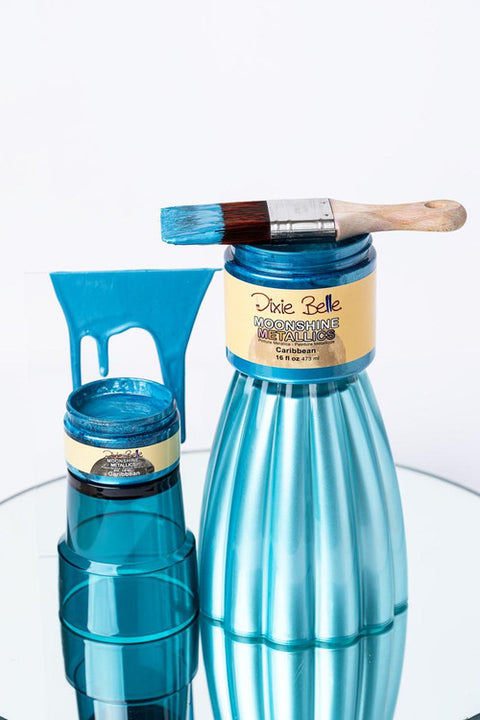 Two blue metallic paint jars, one large and one small, labeled "Dixie Belle Moonshine Metallics Caribbean," are on a mirrored surface. A paintbrush with blue paint rests on the large jar. Blue paint drips behind them.