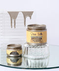 Two jars of Dixie Belle Moonshine Metallics Steel Magnolia paint are displayed on a glass surface, with metallic paint dripping on a clear, ribbed stand in the background. The larger jar is propped up on an inverted glass dish.