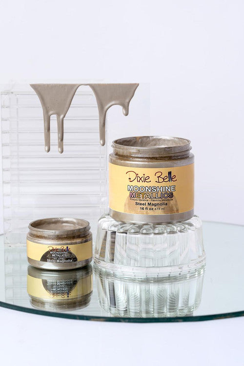 Two jars of Dixie Belle Moonshine Metallics Steel Magnolia paint are displayed on a glass surface, with metallic paint dripping on a clear, ribbed stand in the background. The larger jar is propped up on an inverted glass dish.