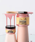 Two jars of Dixie Belle Moonshine Metallics Rozay paint are stacked on pastel pink stands. A paintbrush with pink paint rests on the top jar, with paint dripping down. The scene is set on a reflective surface.