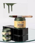 A paintbrush with green paint rests on an open jar of Dixie Belle Moonshine Metallics "Deep Woods" paint, with green drips on a clear panel behind and another open jar below, all on a mirrored surface.