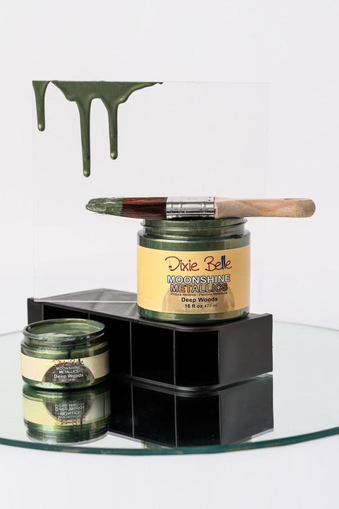 A paintbrush with green paint rests on an open jar of Dixie Belle Moonshine Metallics "Deep Woods" paint, with green drips on a clear panel behind and another open jar below, all on a mirrored surface.