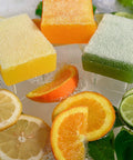 Image of Lemon, Orange, and Lemon Lime Scrubby Soaps out of their packaging, laying next to each other surrounded by lemon, orange, and lime slices, along with leaves and soap suds.