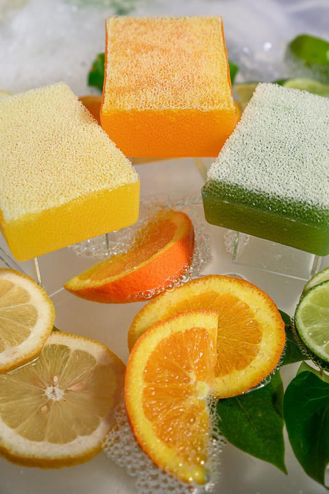 Image of Lemon, Orange, and Lemon Lime Scrubby Soaps out of their packaging, laying next to each other surrounded by lemon, orange, and lime slices, along with leaves and soap suds.