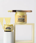 Two jars of Dixie Belle Moonshine Metallics paint in "Wedding Belle" yellow, one open with a paintbrush resting on top and yellow paint dripping, displayed on white and cream blocks.