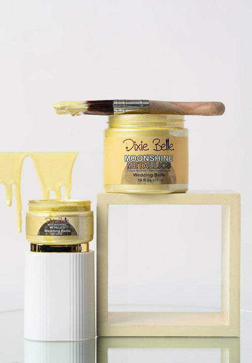 Two jars of Dixie Belle Moonshine Metallics paint in "Wedding Belle" yellow, one open with a paintbrush resting on top and yellow paint dripping, displayed on white and cream blocks.