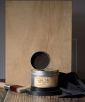 A jar of Dixie Belle Best Dang Wax with an open lid is displayed on a dark circular stand. A paintbrush and black cloth are placed nearby. The background features wooden panels in various shades of brown.