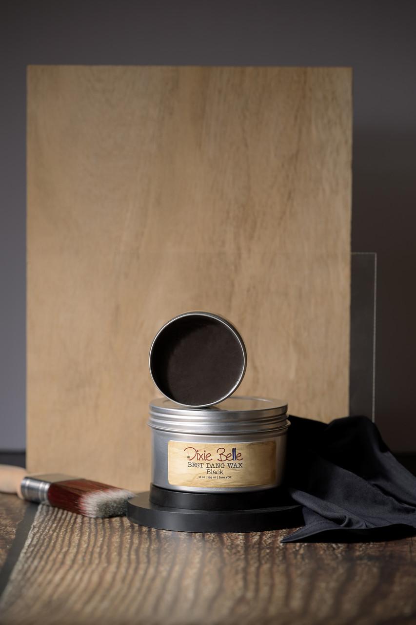 A jar of Dixie Belle Best Dang Wax with an open lid is displayed on a dark circular stand. A paintbrush and black cloth are placed nearby. The background features wooden panels in various shades of brown.