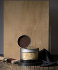 A container of Dixie Belle Best Dang Wax, open to show brown wax inside. It is placed on a wooden surface with a wooden backdrop, accompanied by a paintbrush and black cloth.