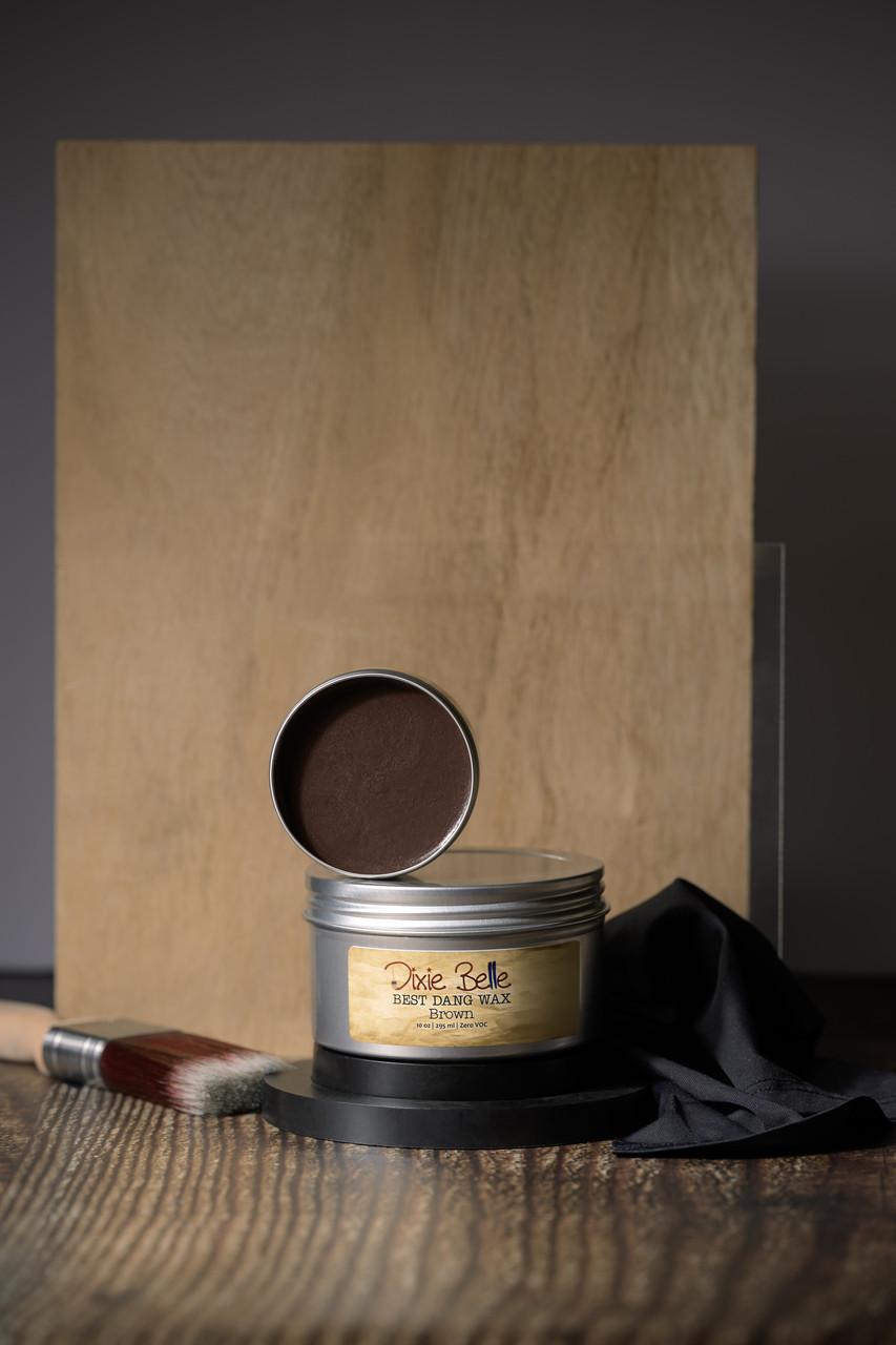 A container of Dixie Belle Best Dang Wax, open to show brown wax inside. It is placed on a wooden surface with a wooden backdrop, accompanied by a paintbrush and black cloth.