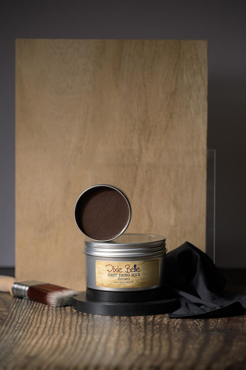 A container of Dixie Belle Best Dang Wax, open to show brown wax inside. It is placed on a wooden surface with a wooden backdrop, accompanied by a paintbrush and black cloth.