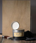 A tin of Dixie Belle Best Dang Wax with the lid off, displaying the clear wax inside. It is placed on a wooden surface with a painter's brush and dark cloth nearby. The background features a wooden board.