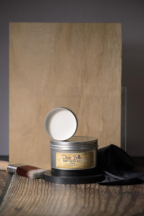 A tin of Dixie Belle Best Dang Wax with the lid off, displaying the clear wax inside. It is placed on a wooden surface with a painter's brush and dark cloth nearby. The background features a wooden board.
