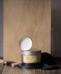 A container of Dixie Belle Best Dang Wax in white is open, resting on top of a 10 oz. jar. A paintbrush and black cloth are nearby. The items are set against a wooden board background.