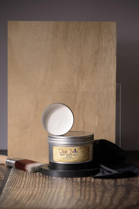 A container of Dixie Belle Best Dang Wax in white is open, resting on top of a 10 oz. jar. A paintbrush and black cloth are nearby. The items are set against a wooden board background.