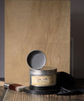 A jar of Dixie Belle Best Dang Wax stands open on a wood surface, revealing a light gray wax inside. A paintbrush and a black cloth are placed next to it, with two pieces of wood in the background.