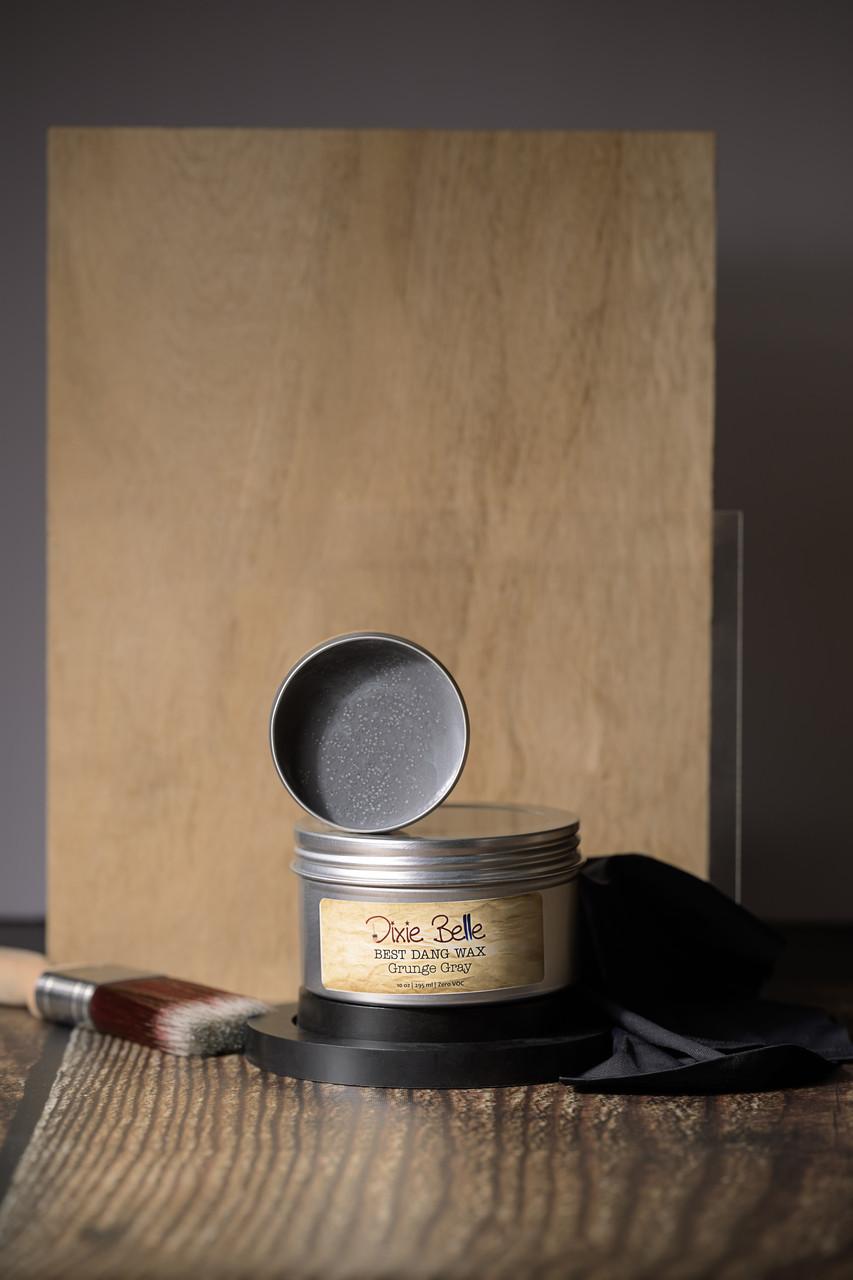 A jar of Dixie Belle Best Dang Wax stands open on a wood surface, revealing a light gray wax inside. A paintbrush and a black cloth are placed next to it, with two pieces of wood in the background.