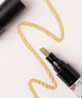 A black marker with a gold glitter tip is shown with the cap off. The marker is drawing a wavy gold line on textured white paper. The design on the marker barrel includes floral patterns.