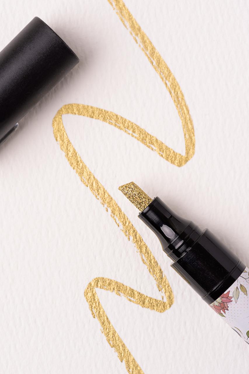 A black marker with a gold glitter tip is shown with the cap off. The marker is drawing a wavy gold line on textured white paper. The design on the marker barrel includes floral patterns.