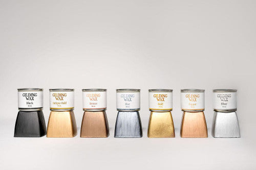 Seven jars of Gilding Wax in different colors—black, antique gold, bronze, zinc, gold, copper, and silver—are lined up on a white background, each shown above a swatch of its corresponding color.