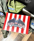 A painted cigar box with red and white stripes, with an eagle in front of American flags and "God Bless America" on the lid is placed on the floor. Surrounding the box are military-style jackets in green with patches, one reading "BELLION," and another displaying an emblem.