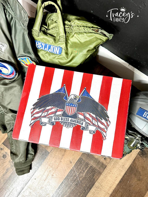 A painted cigar box with red and white stripes, with an eagle in front of American flags and "God Bless America" on the lid is placed on the floor. Surrounding the box are military-style jackets in green with patches, one reading "BELLION," and another displaying an emblem.