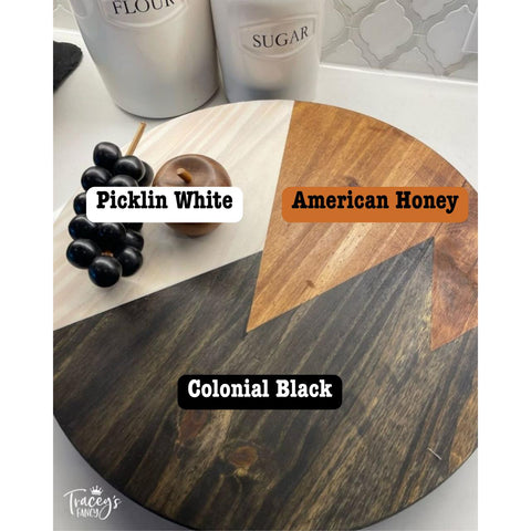 A round wooden board shows three sections with different wood stains: Picklin White, American Honey, and Colonial Black. A cup of black grapes and a small wooden apple rest on the board. Containers labeled Flour and Sugar are in the background.