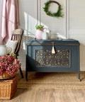 A dark teal storage chest with floral paneling stands against white shutters. It's topped with a white pitcher and pink teapot. A wicker basket with red hydrangeas and an armchair are on the left. A green wreath hangs above. Warm, cozy setting.