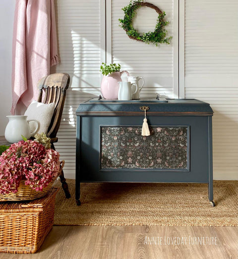 A dark teal storage chest with floral paneling stands against white shutters. It's topped with a white pitcher and pink teapot. A wicker basket with red hydrangeas and an armchair are on the left. A green wreath hangs above. Warm, cozy setting.