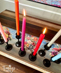 A set of six colorful candles in black candle holders are arranged on a wooden surface with floral designs. The candles are in shades of orange, pink, red, blue and white, with three of them lit. "Tracey's Fancy" is written on the bottom left corner of the image.