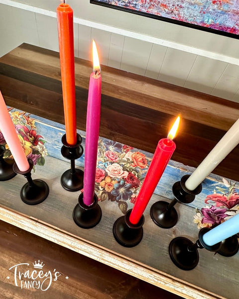 A set of six colorful candles in black candle holders are arranged on a wooden surface with floral designs. The candles are in shades of orange, pink, red, blue and white, with three of them lit. "Tracey's Fancy" is written on the bottom left corner of the image.