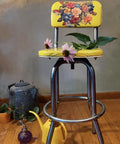 Yellow barstool with a floral pattern on the backrest, placed on a wooden floor against a muted wall. On the seat are two pink flowers and leaves. Nearby are a blue teapot, a yellow watering can, a purple decorative glass, and potted plants. "MTM artistry" watermark logo on the floor.