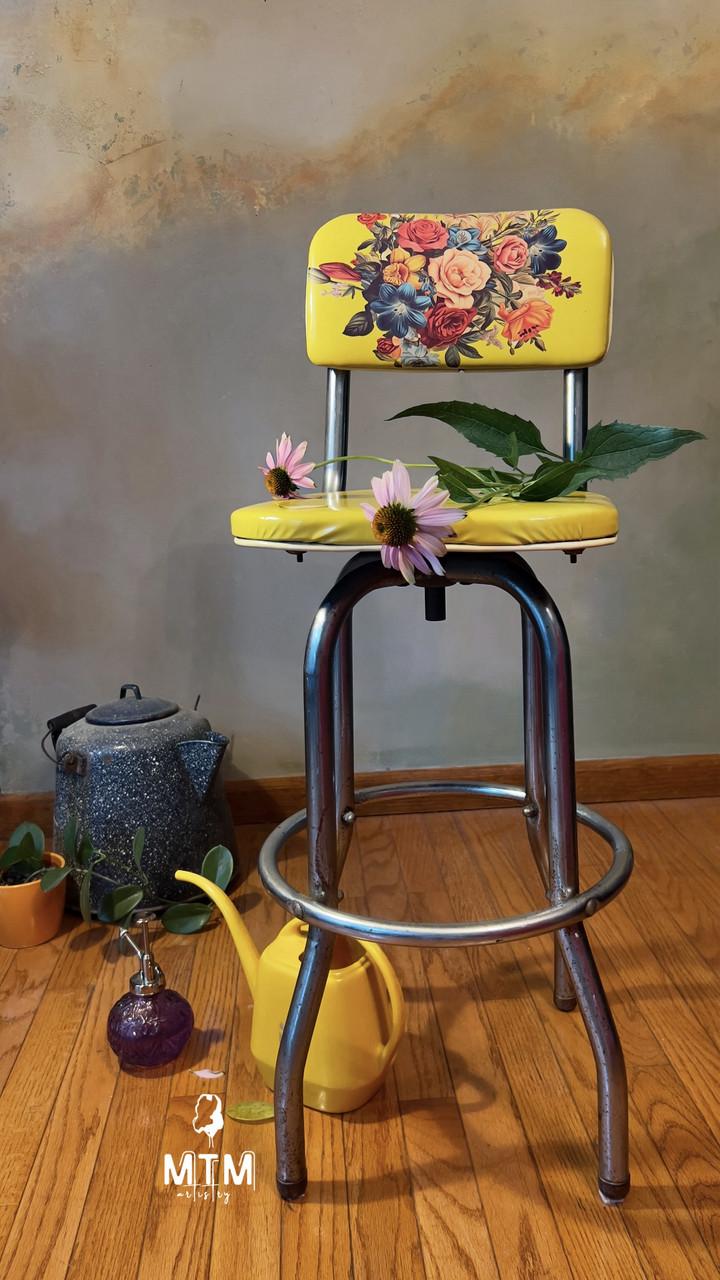 Yellow barstool with a floral pattern on the backrest, placed on a wooden floor against a muted wall. On the seat are two pink flowers and leaves. Nearby are a blue teapot, a yellow watering can, a purple decorative glass, and potted plants. "MTM artistry" watermark logo on the floor.