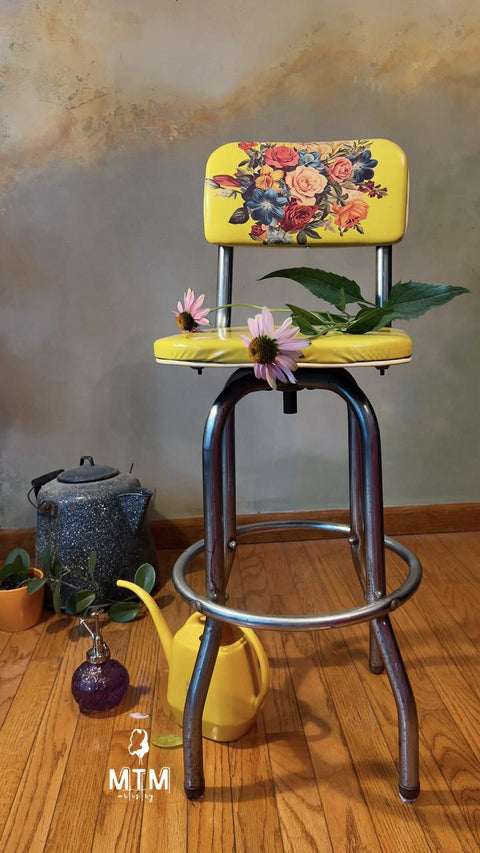 Yellow barstool with a floral pattern on the backrest, placed on a wooden floor against a muted wall. On the seat are two pink flowers and leaves. Nearby are a blue teapot, a yellow watering can, a purple decorative glass, and potted plants. "MTM artistry" watermark logo on the floor.