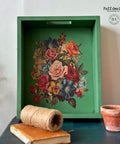 A green wooden tray adorned with a vibrant floral design is positioned upright against a wall. In front of it, on a black surface, there is a spool of twine, a small clay pot, an orange candle, and a closed book. "Faff designs" watermark logo is placed on the wall.