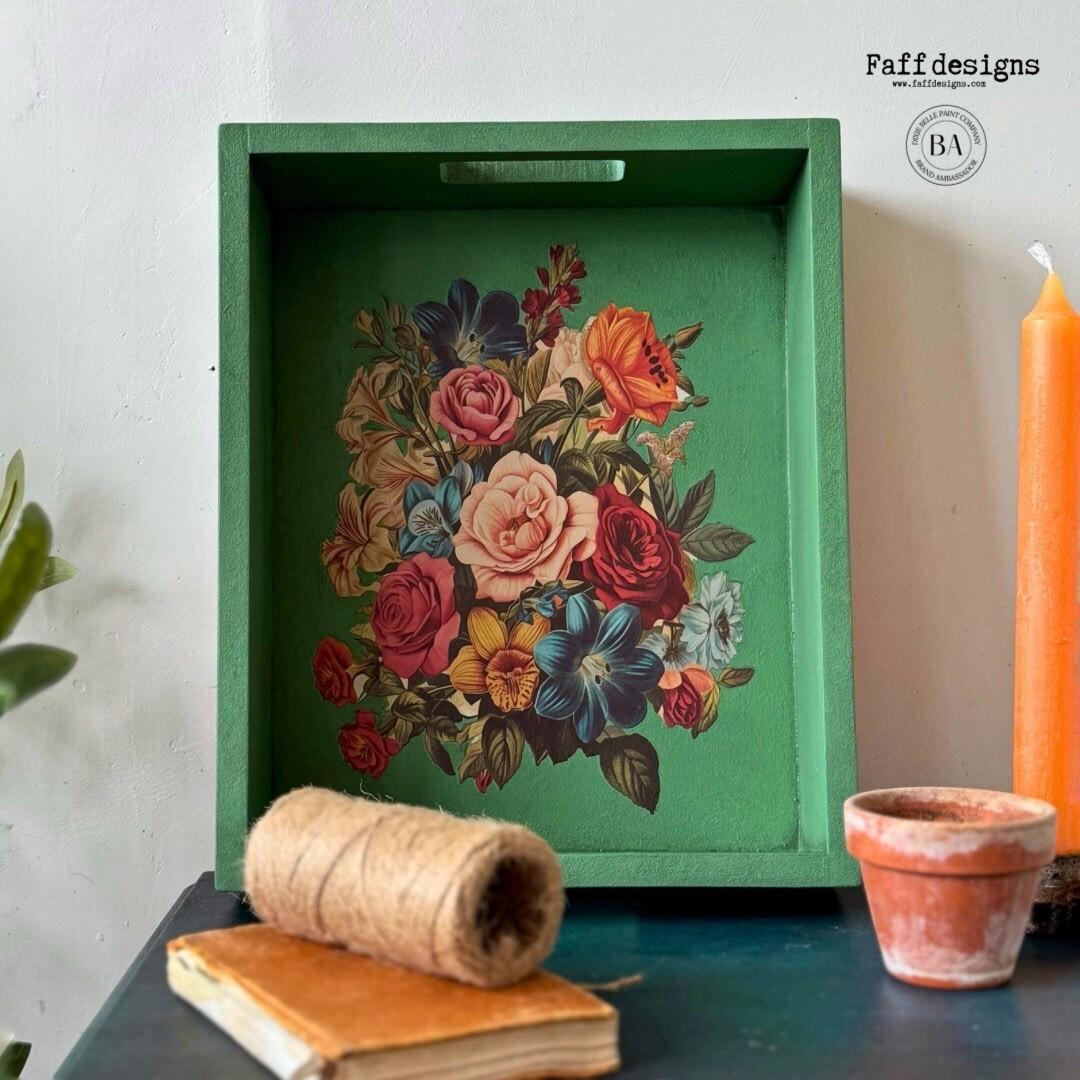 A green wooden tray adorned with a vibrant floral design is positioned upright against a wall. In front of it, on a black surface, there is a spool of twine, a small clay pot, an orange candle, and a closed book. "Faff designs" watermark logo is placed on the wall.