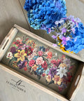 A wooden tray on a table features a vibrant array of flowers, including roses, lilies, and other blooms. Next to the tray, a bouquet of blue and purple hydrangeas adds a touch of freshness. "Leah Kendrick Design" watermark logo placed below the tray.