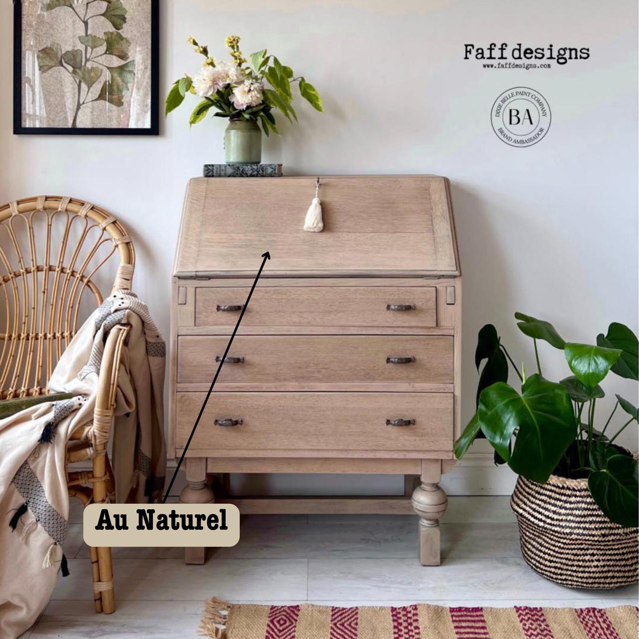 A cozy room featuring a light wood writing desk with a vase of flowers on top. A wicker chair with a blanket sits beside it. A decorative rug is on the floor, with a basket of leafy green plants nearby. Text reads "Au Naturel."