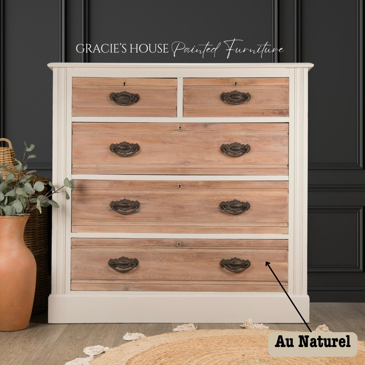 A wooden dresser with a light cream frame and natural wood drawers, featuring ornate metal handles. It's set against a dark paneled wall, accompanied by a basket with a plant and a terracotta pot on a light rug. A label reads "Au Naturel."