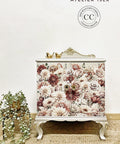 A small vintage-style cabinet adorned with a floral print featuring roses and peonies. The cabinet stands on ornate legs and has a gold decorative trim on top. Two gold bird ornaments sit atop it. A leafy plant is placed to the left of the cabinet.