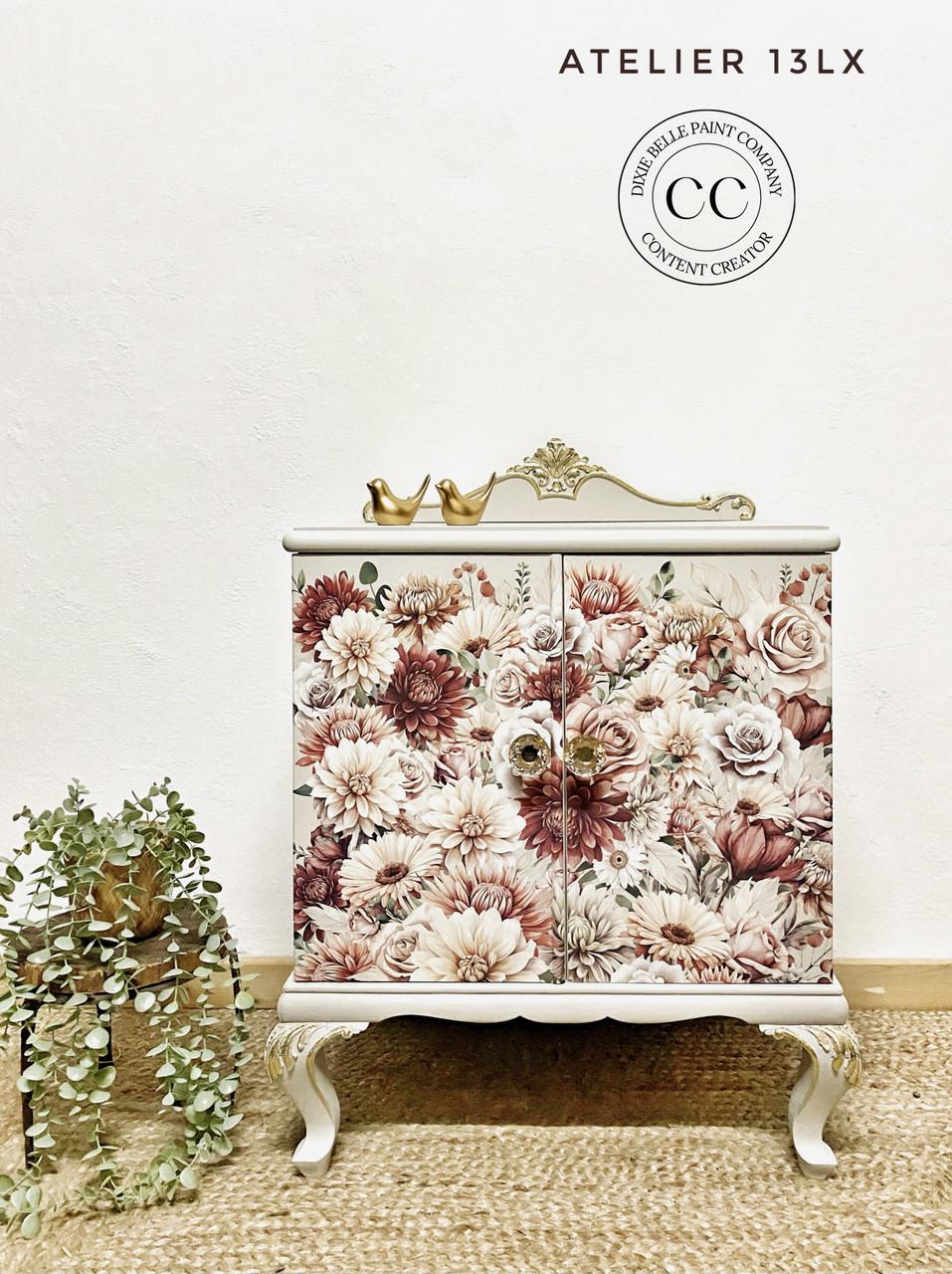 A small vintage-style cabinet adorned with a floral print featuring roses and peonies. The cabinet stands on ornate legs and has a gold decorative trim on top. Two gold bird ornaments sit atop it. A leafy plant is placed to the left of the cabinet.