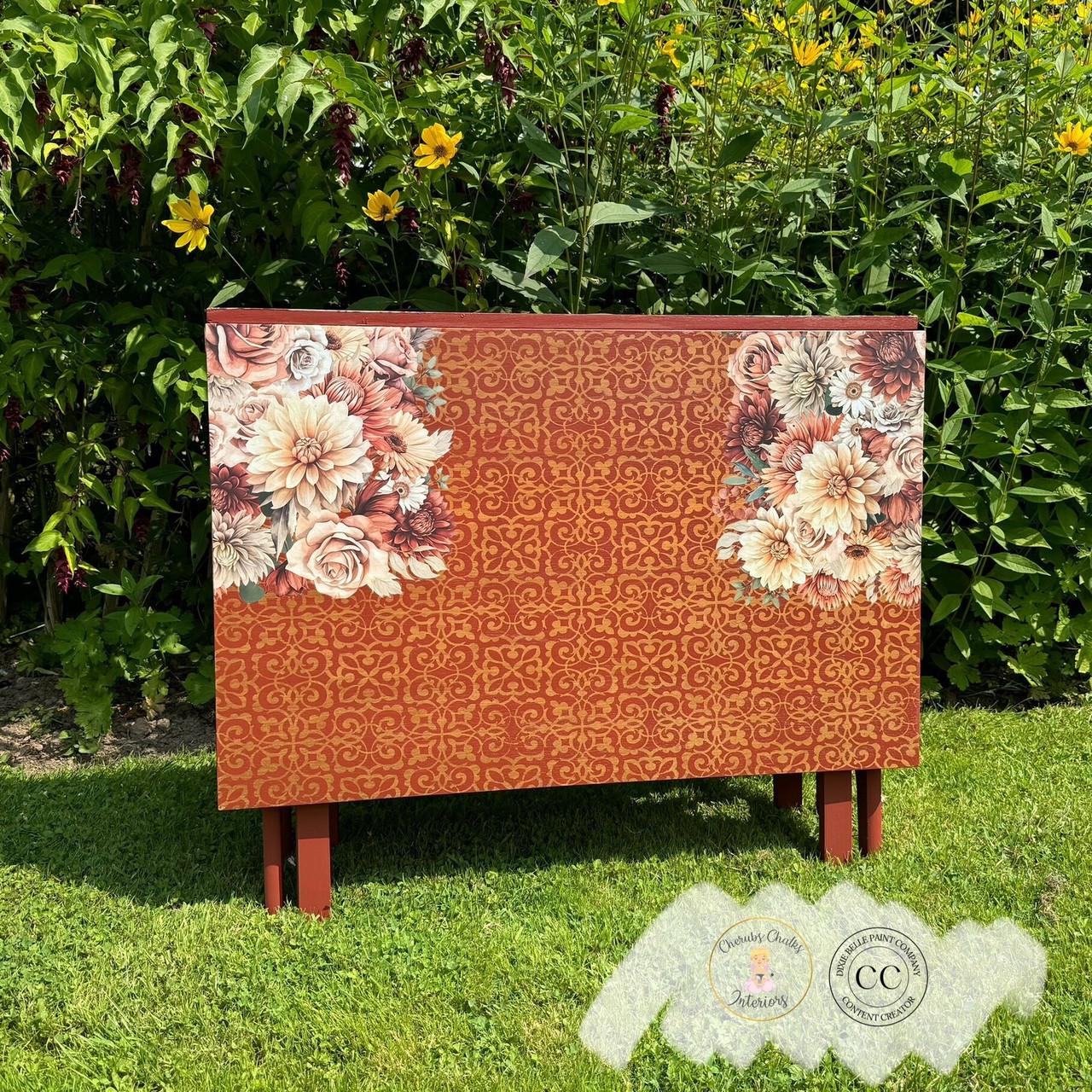 A wooden chest sits on a grassy lawn in front of tall green plants and yellow flowers. The chest is decorated with intricate patterns in red and gold, and features floral designs of large blossoms on its top corners. Two small certification badges are visible at the bottom.