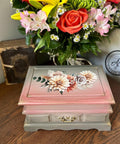 A vintage wooden jewelry box with a floral design on the top sits on a wooden table. Behind the box, there's a large bouquet of colorful flowers in a variety of colors and a worn book. A watermark in the corner reads "AJ's Vintage Designs"