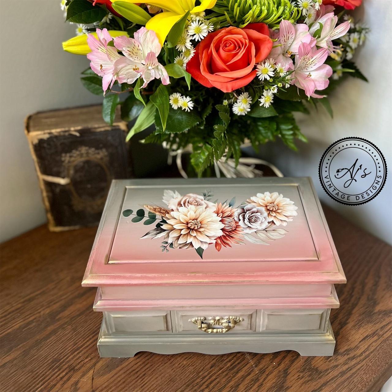 A vintage wooden jewelry box with a floral design on the top sits on a wooden table. Behind the box, there's a large bouquet of colorful flowers in a variety of colors and a worn book. A watermark in the corner reads "AJ's Vintage Designs"