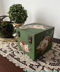 A small, square, green wooden box with metal latches, adorned with floral designs. The box is placed on a lace tablecloth, accompanied by a decorative green ball of faux foliage and a bronze decorative object. Watermark reads "Creative Moments"